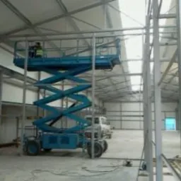 Montaż konstrukcji stalowej hali z użyciem podnośnika nożycowego, widoczne elementy ramy i oświetlenie hali w tle.