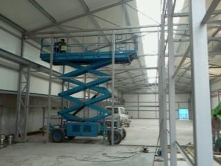 Montaż konstrukcji stalowej hali z użyciem podnośnika nożycowego, widoczne elementy ramy i oświetlenie hali w tle.