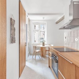 Browwar Nieruchomości - agent Michał Zwierz - Jasna kuchnia z drewnianymi meblami, okrągłym stołem przy oknie i dekoracyjną makramą na ścianie.