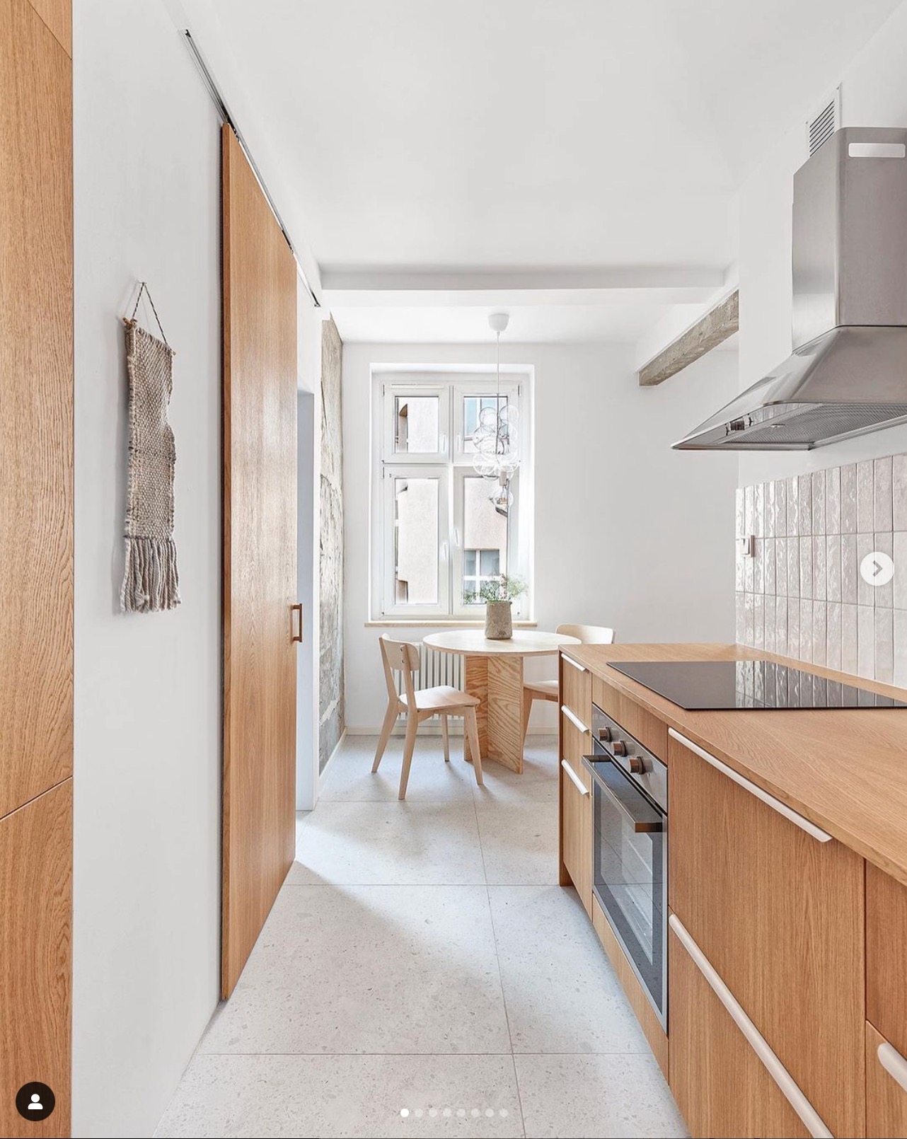 Jasna kuchnia z drewnianymi meblami, okrągłym stołem przy oknie i dekoracyjną makramą na ścianie.