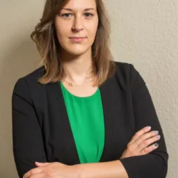 Portret kobiety w czarnej marynarce i zielonej bluzce, z założonymi rękami na tle kremowej ściany, z delikatnym makijażem i minimalistyczną biżuterią.