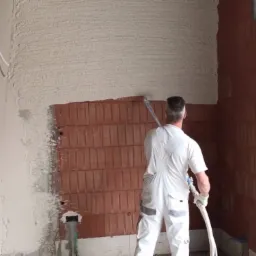 Pracownik w białym kombinezonie nanosi tynk maszynowy na ceglaną ścianę w surowym stanie, widoczne przewody elektryczne i profile okienne.