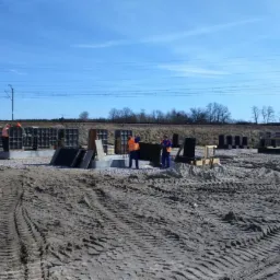 Budowa fundamentów pod konstrukcję, widoczni pracownicy w odblaskowych kamizelkach, szalunki zbrojeniowe, teren budowy z widocznymi śladami opon i wałem ziemi w tle.