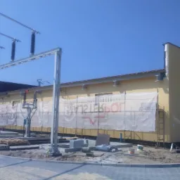 Budynek stacji transformatorowej w trakcie budowy, z widocznymi elementami konstrukcji stalowych, osłonami i przygotowanym terenem.