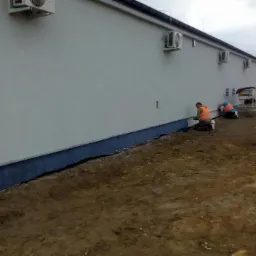 Pracownicy w pomarańczowych kamizelkach i kaskach malują dolną część elewacji budynku na ciemno niebiesko, widoczne klimatyzatory na ścianie, teren wokół w trakcie prac ziemnych.