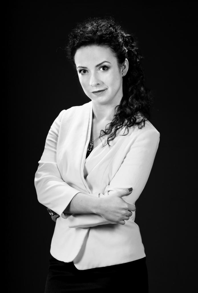 Portret kobiety w białym żakiecie z założonymi rękami, na czarnym tle, czarno-białe zdjęcie.