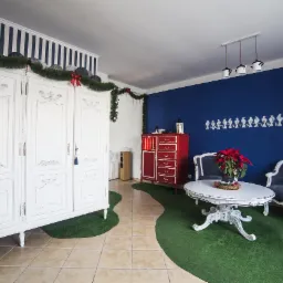 Udekorowane wnętrze pokoju z białymi meblami w stylu vintage, czerwonymi akcentami i sztuczną trawą na podłodze.