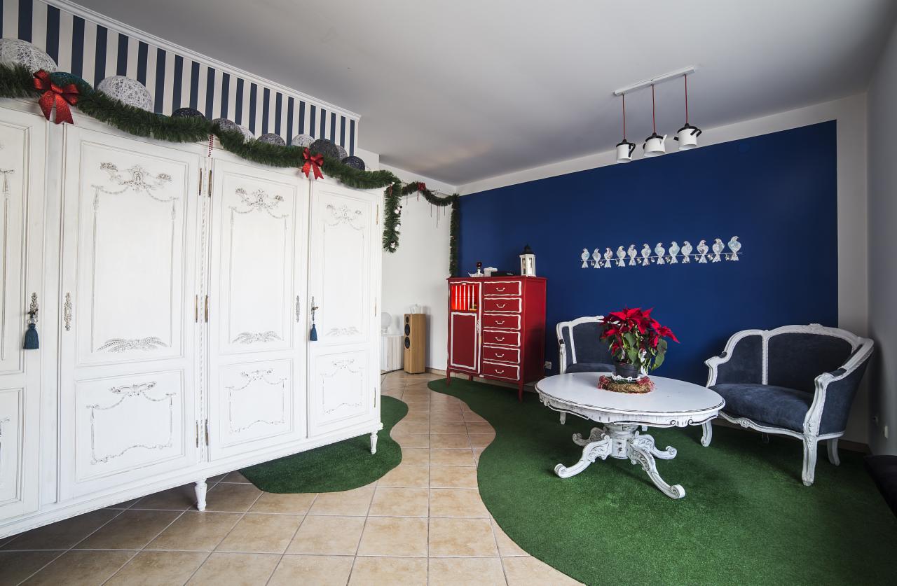 Udekorowane wnętrze pokoju z białymi meblami w stylu vintage, czerwonymi akcentami i sztuczną trawą na podłodze.