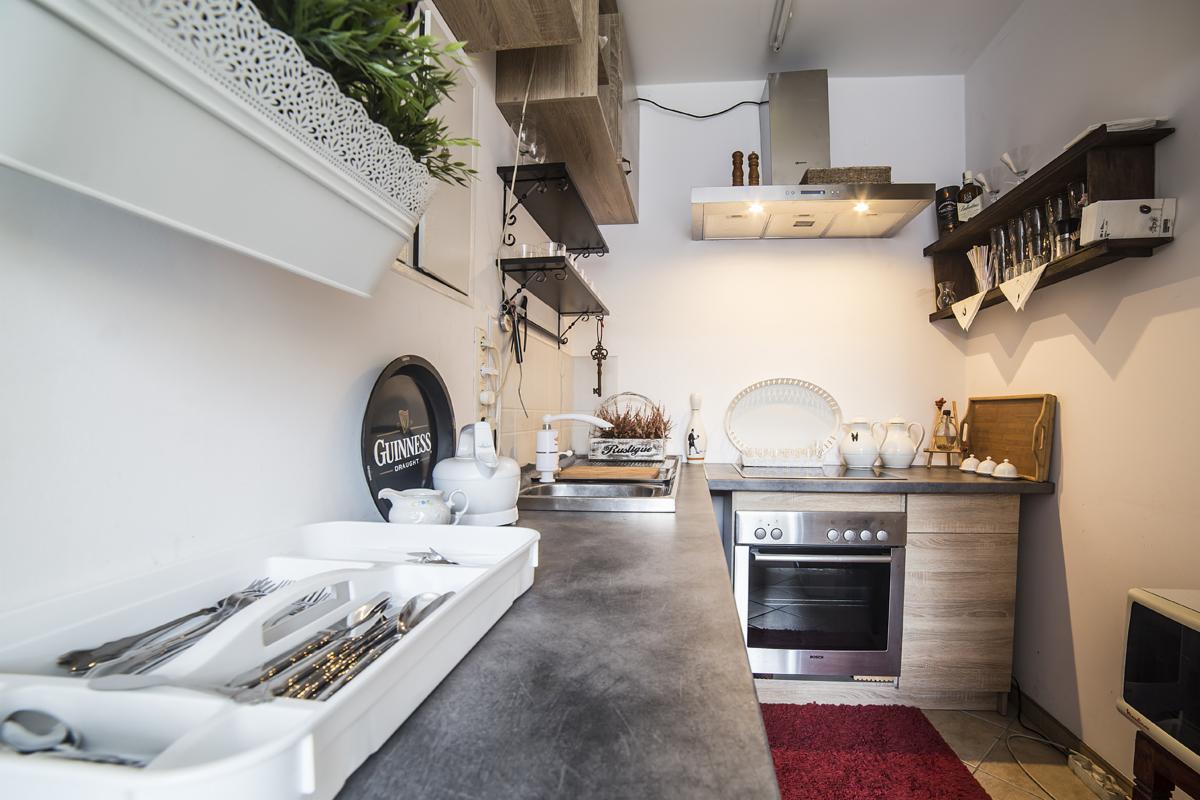 Wnętrze kuchni z szarym blatem, stalowym piekarnikiem i drewnianymi szafkami, nad zlewem wiszą półki z dekoracjami, a na ścianie widoczny jest szyld Guinness