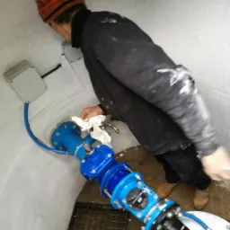 Instalacja wodociągowa w studni głębinowej w Maszewie: pracownik czyści złącza niebieskich rur, widoczna skrzynka elektryczna i przewód.