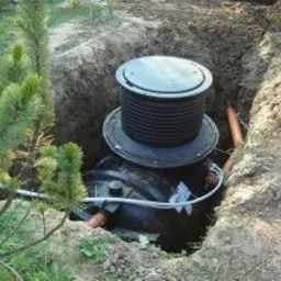 Osadzony w ziemi zbiornik z czarnym, żebrowanym wierzchem i białym przewodem, widoczny w wykopie otoczonym ziemią i roślinnością.