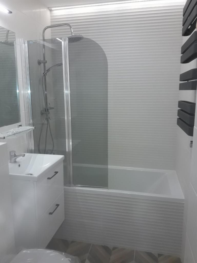 Nowoczesna, biała łazienka z wanną z parawanem nawannowym, umywalką z szafką i grzejnikiem drabinkowym, wykończona płytkami z poziomym wzorem i podłogą z płytek w jodełkę.