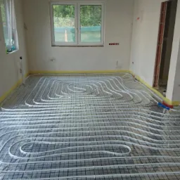 Białe rury ogrzewania podłogowego ułożone na siatce montażowej w surowym pomieszczeniu z oknami i żółtą taśmą maskującą przy ścianach.