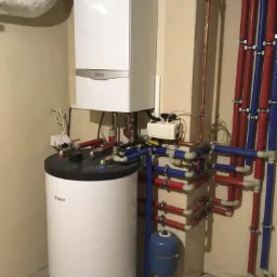 Biały piec gazowy wiszący nad zasobnikiem ciepłej wody, z widocznymi rurami w izolacji czerwonej i niebieskiej oraz małym niebieskim zbiornikiem wyr&oacute;wnawczym, całość zamontowana w rogu pomieszczenia.