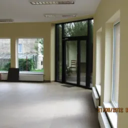 Wnętrze pustego pomieszczenia z dużymi oknami i przeszklonymi drzwiami, widoczne grzejniki i krzesło w tle, ściany w odcieniach beżu.