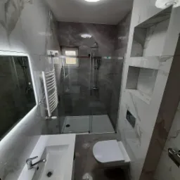 Nowoczesna łazienka z kabiną prysznicową ze szklanymi drzwiami, umywalką z lustrem podświetlanym LED, grzejnikiem drabinkowym i toaletą w odcieniach szarości i bieli, wykończona płytkami...