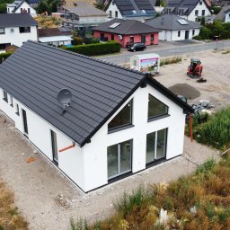 WOOD- HOME SL&S AG Sp. z o.o. - Nowoczesny, biały dom z czarnym dachem w trakcie budowy, widziany z góry. Na dachu antena satelitarna, w tle inne domy i plac budowy z mini koparką.