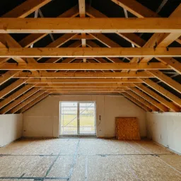 Wnętrze domu w stanie surowym zamkniętym: drewniana konstrukcja dachu, okno z widokiem na zieleń, podłoga z płyt OSB. Widoczne elementy instalacji.