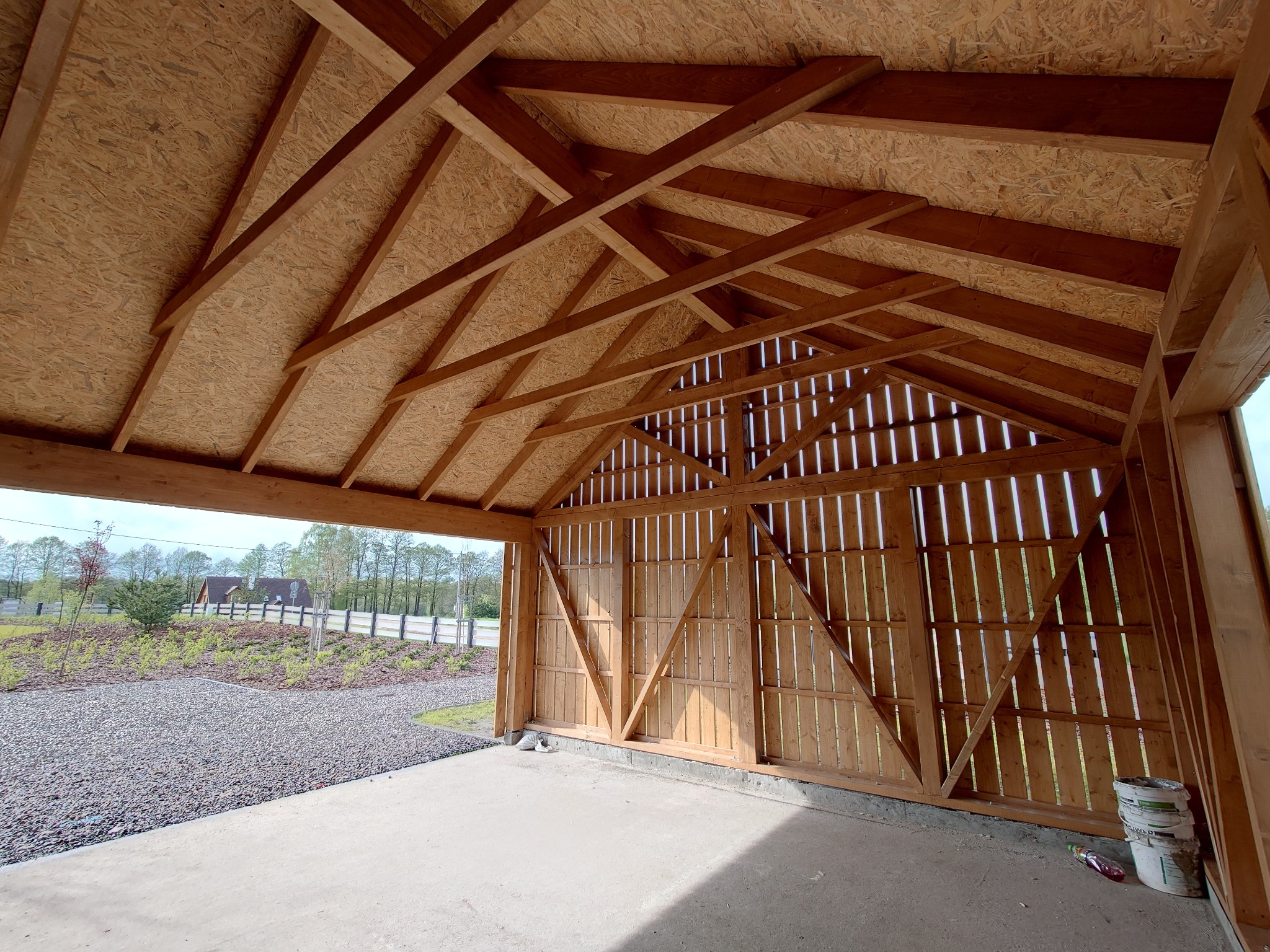 Wnętrze garażu szkieletowego w trakcie budowy, widoczna drewniana konstrukcja dachu z poszyciem z płyty OSB, ściany z pionowych desek z prześwitami, betonowa posadzka i widok na ogród z zewnątrz.