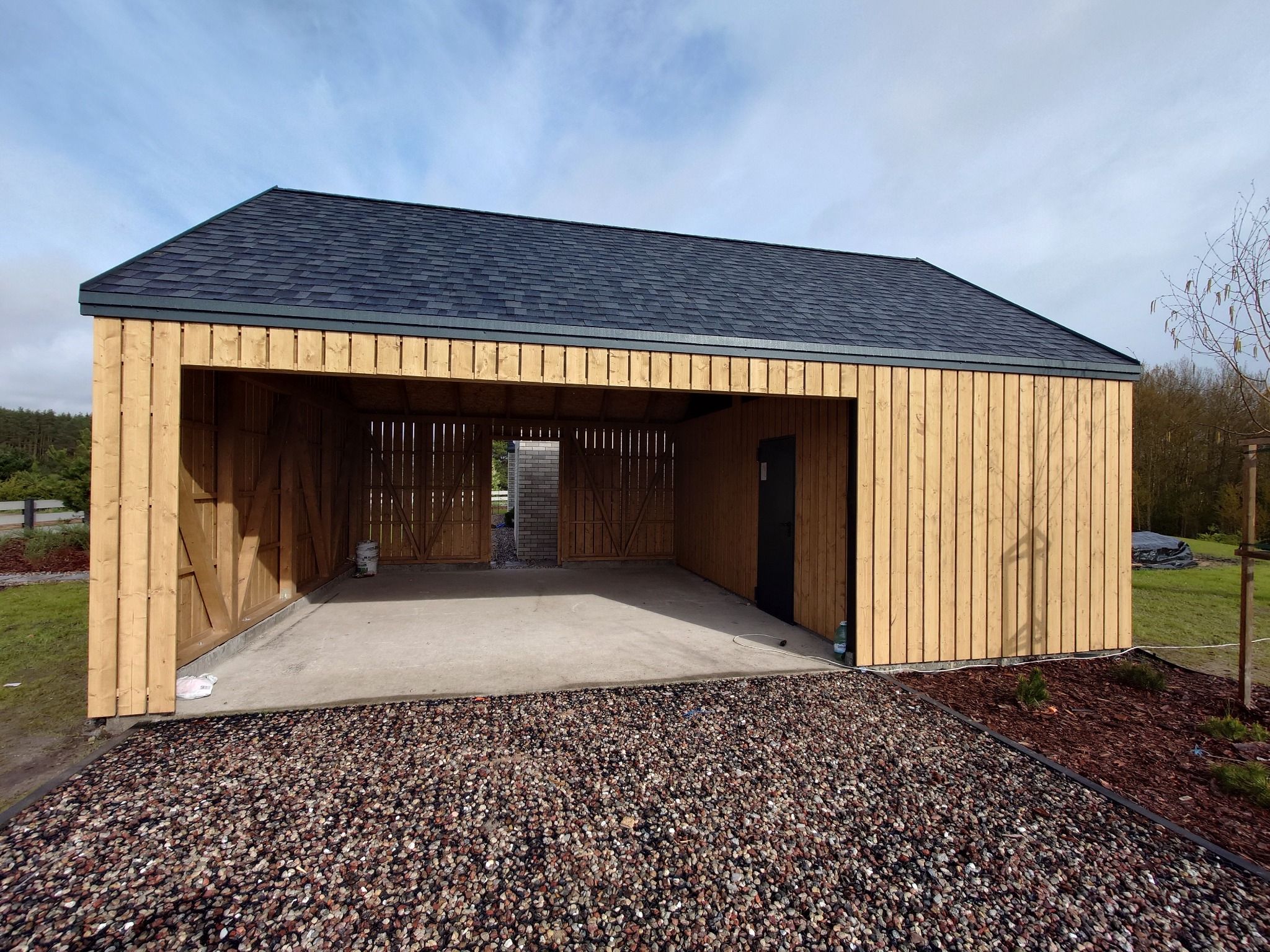 Nowoczesny, otwarty garaż z drewna z szarą dachówką bitumiczną, wykończony kamiennym podłożem i elementami zagospodarowania terenu.