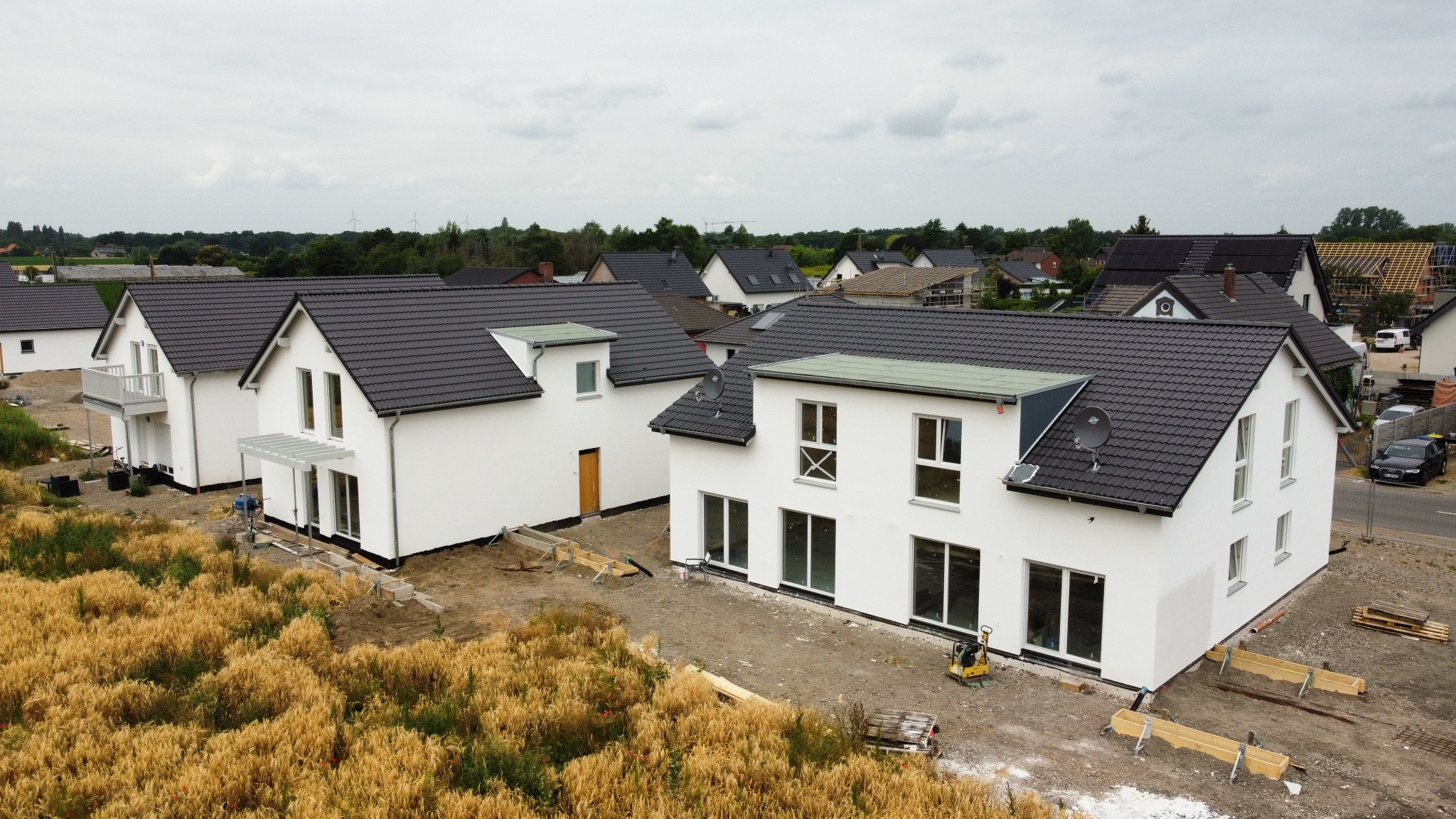 Nowoczesne, białe domy z ciemnymi dachami w zabudowie szeregowej, widoczne z góry, otoczone polami i innymi domami, w trakcie prac wykończeniowych.
