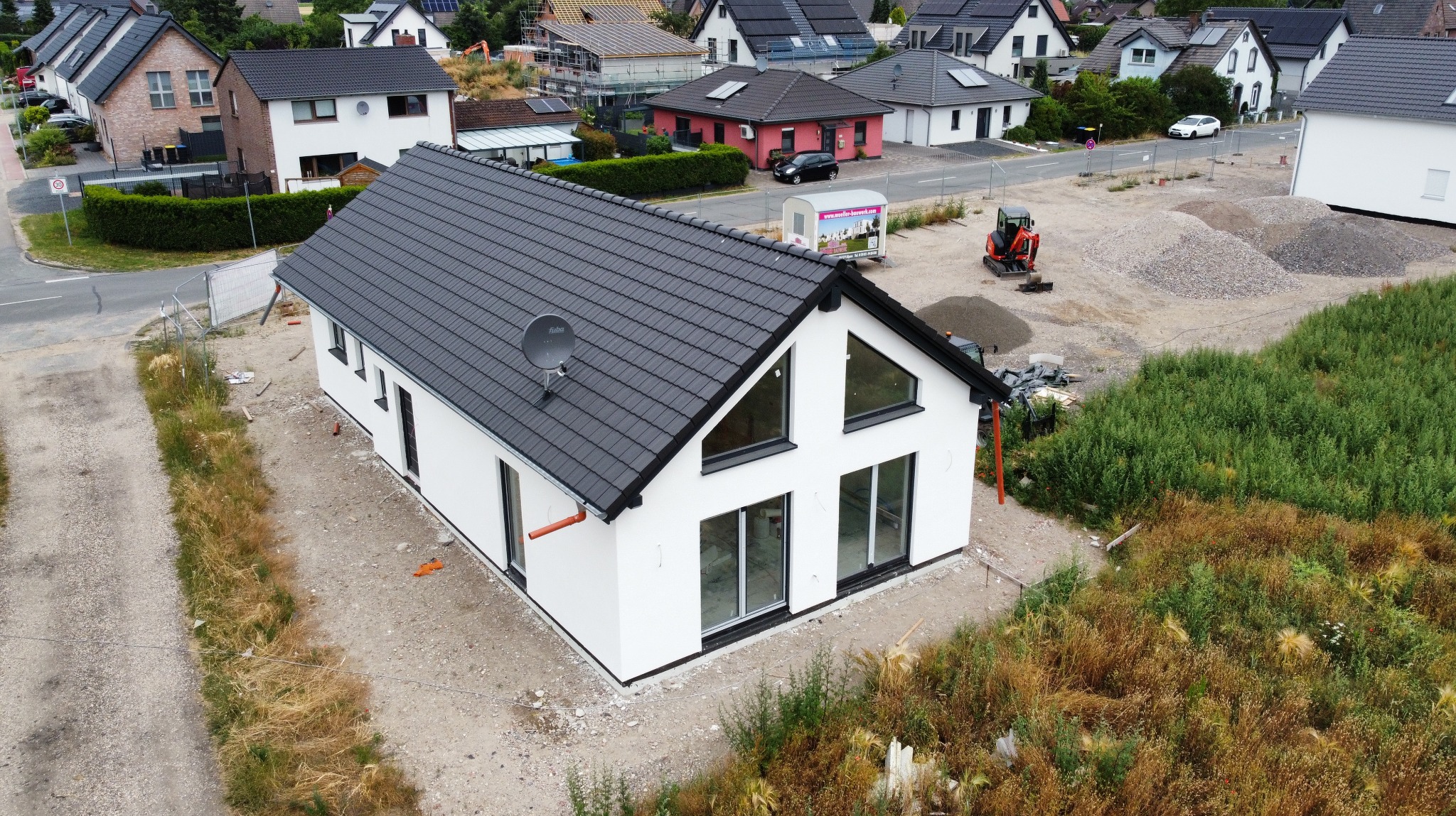 Nowoczesny, biały dom z czarnym dachem w trakcie budowy, widziany z góry. Na dachu antena satelitarna, w tle inne domy i plac budowy z mini koparką.
