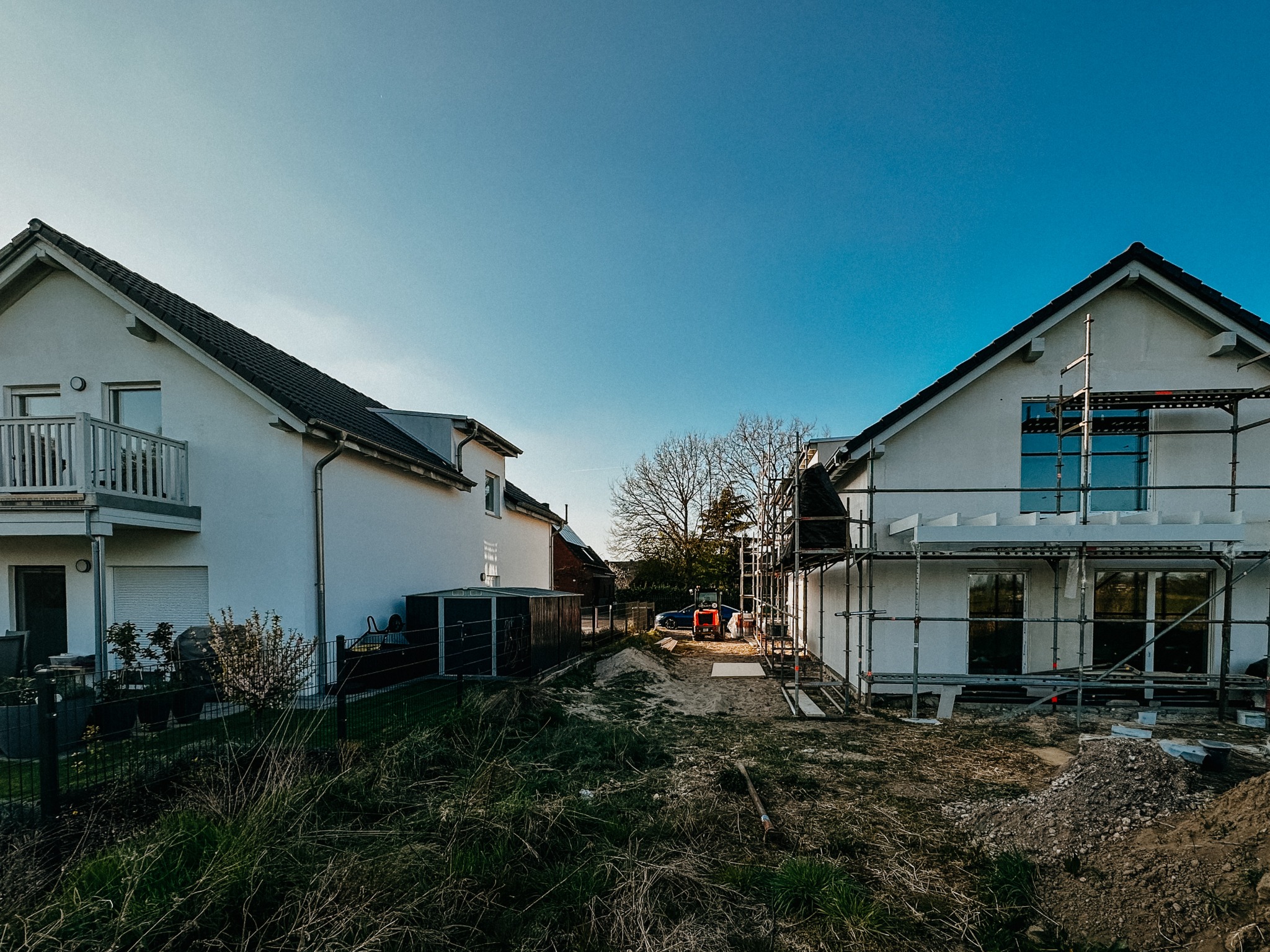 Budowa domu: białe budynki w zabudowie bliźniaczej, jeden ze rusztowaniem, w tle błękitne niebo i drzewa. Widok z poziomu gruntu na plac budowy.