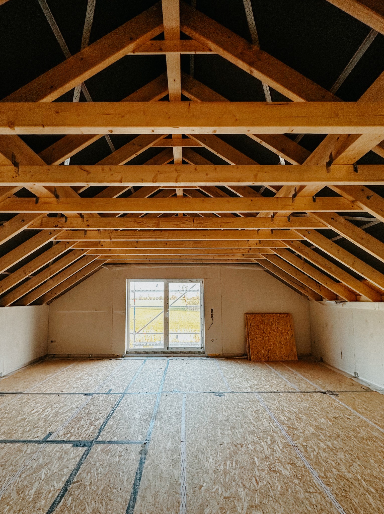 Wnętrze domu w stanie surowym zamkniętym: drewniana konstrukcja dachu, okno z widokiem na zieleń, podłoga z płyt OSB. Widoczne elementy instalacji.