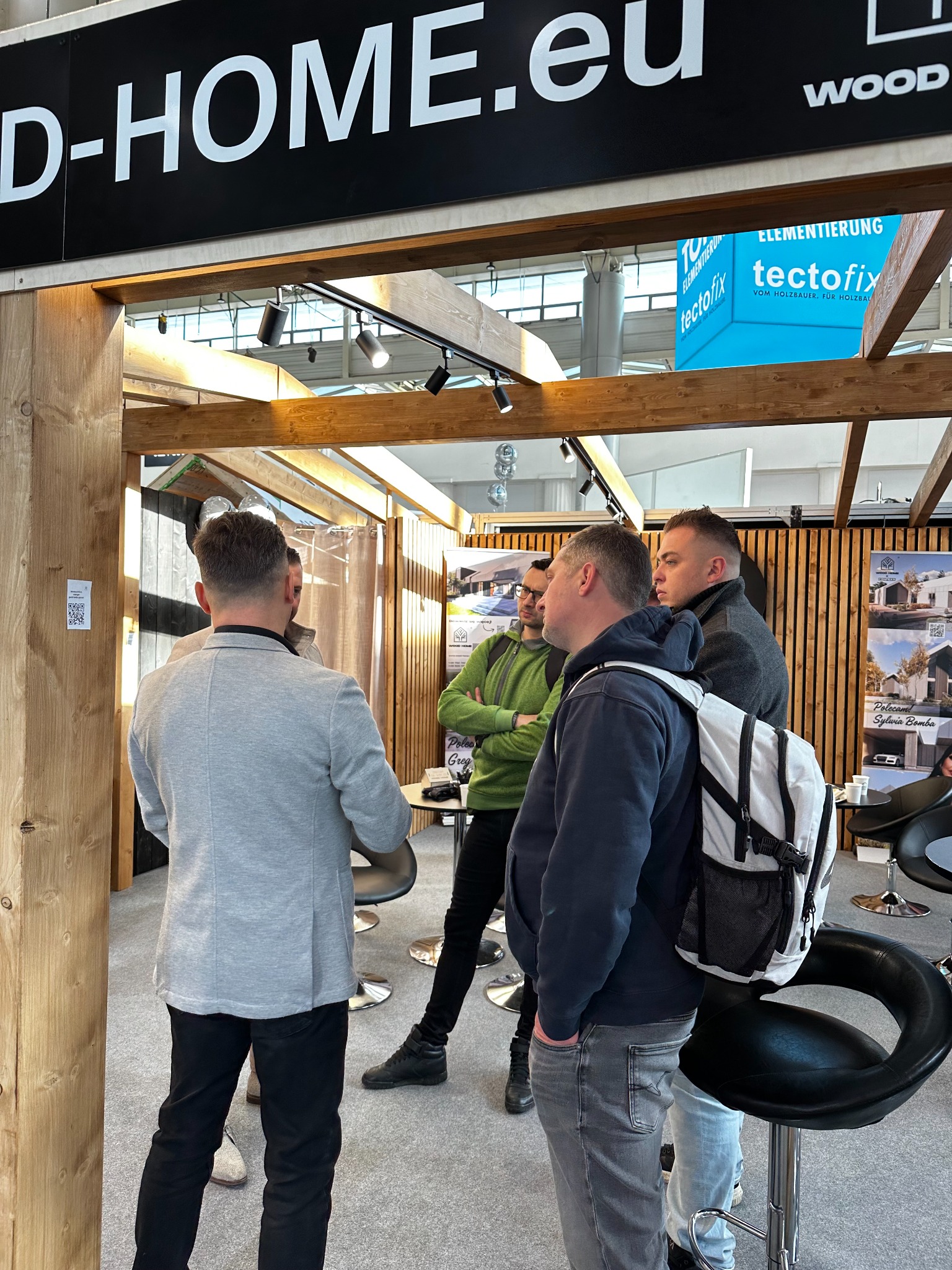 Wnętrze stoiska targowego z drewnianą konstrukcją, logo 'D-HOME.eu WOOD' i grupą osób rozmawiających. Widoczne plakaty z projektami domów.