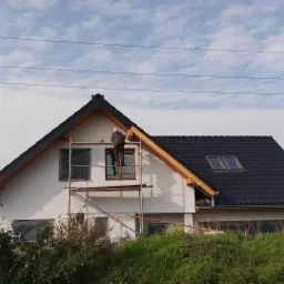 Pracownik na rusztowaniu przy białym domu z czarnym dachem, widoczne drewniane elementy konstrukcji dachu i okna zabezpieczone folią.