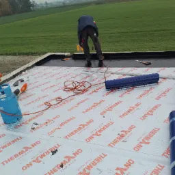 Praca dekarza na dachu płaskim: układanie papy termozgrzewalnej marki BMI Icopal, widoczny palnik gazowy, rolki papy i narzędzia, w tle zielone pole.