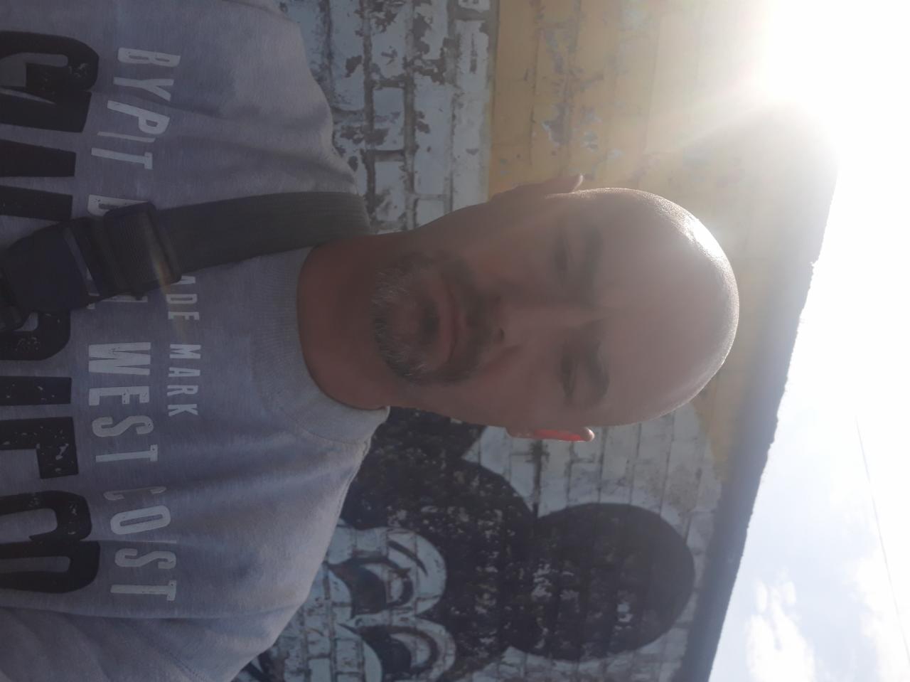 Mężczyzna z brodą i szelką na tle ściany z graffiti i łuszczącej się farby, oświetlonej mocnym słońcem. Nietypowe ujęcie.