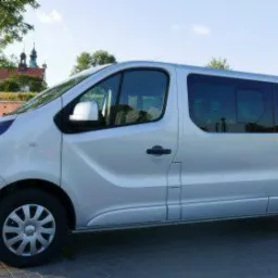 Srebrny bus Opel Vivaro z przyciemnianymi szybami na tle ceglanego muru i kościoła z wieżami.