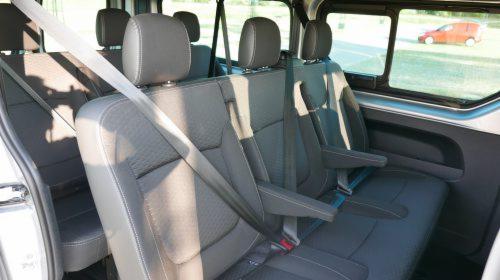 Wnętrze busa z rzędami szarych siedzeń, widok z boku na pasy bezpieczeństwa i zagłówki, czerwony samochód widoczny przez okno.