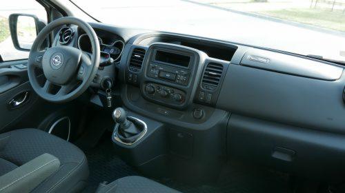 Wnętrze samochodu Opel Vivaro z widoczną kierownicą z logo, deską rozdzielczą i dźwignią zmiany biegów, z fotelami obitymi ciemną tapicerką.