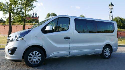 Srebrny bus Opel Vivaro z przyciemnianymi szybami na tle ceglanego muru i kościoła z wieżami.