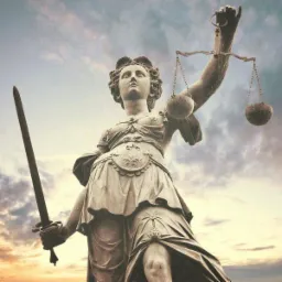 Pomnik Temidy, bogini sprawiedliwości, z mieczem i wagą na tle pochmurnego nieba o zmierzchu, widziany z dołu.