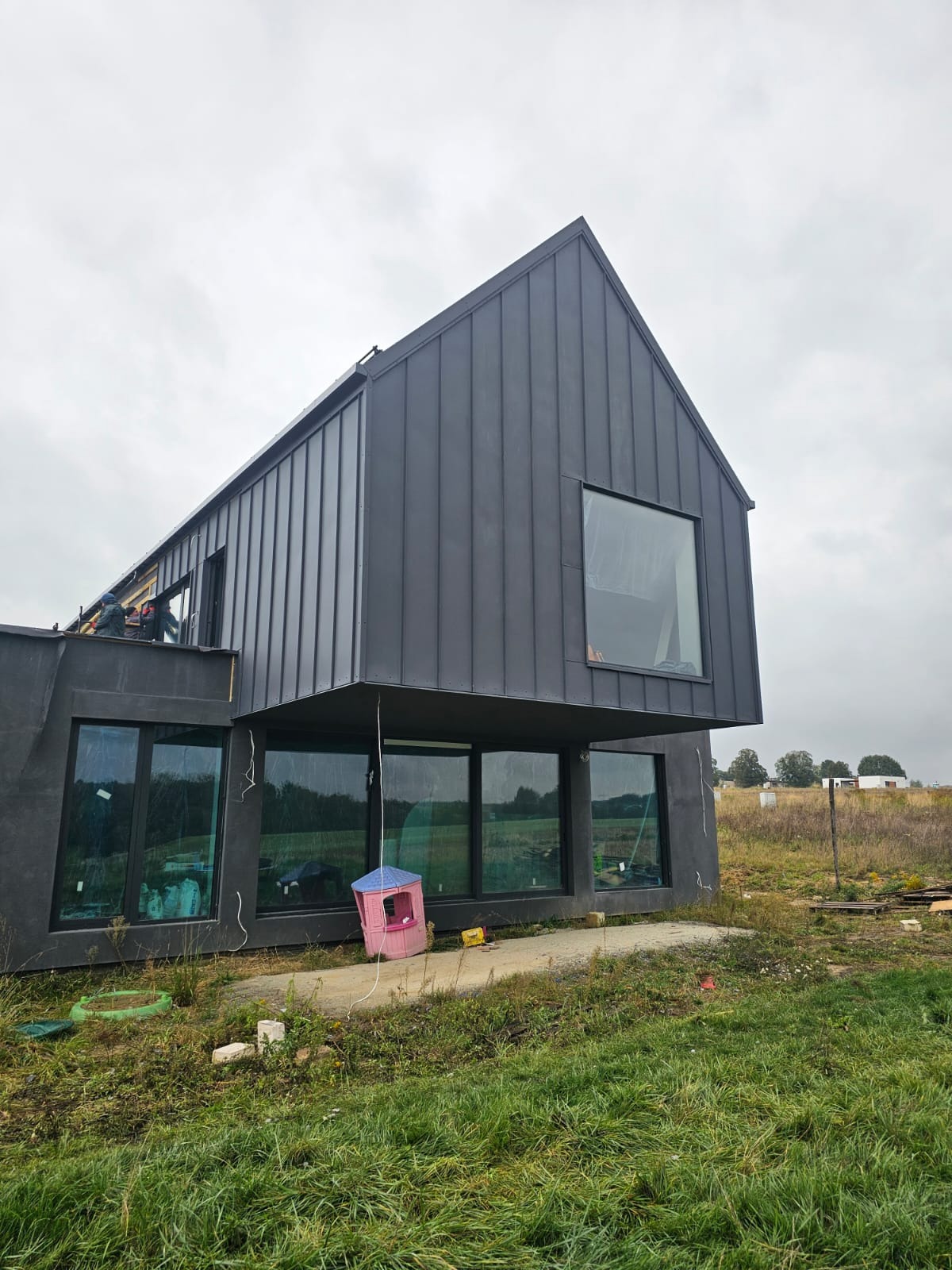 Surowy, nowoczesny dom z ciemną elewacją w Gliwicach, w trakcie budowy, z widocznym fragmentem dachu i dużymi oknami, otoczony polną roślinnością i z małym, plastikowym domkiem dla dzieci w pobliżu.