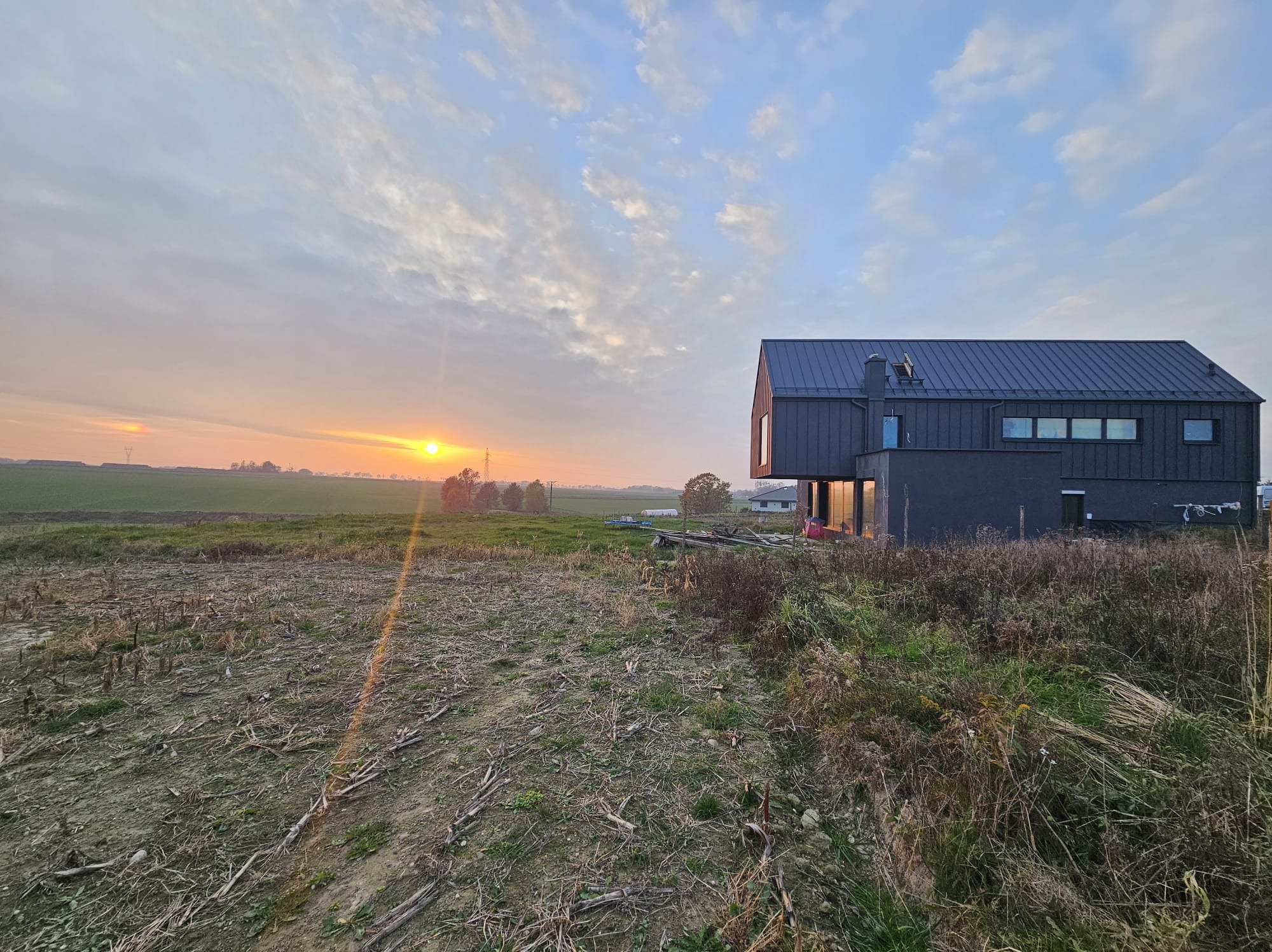 Nowoczesny dom z ciemną elewacją i metalowym dachem o zachodzie słońca, widok na pole i odległe budynki.