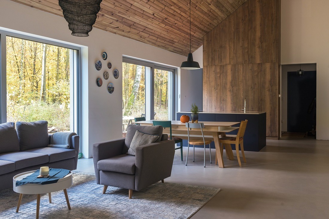 Jasne wnętrze domu z drewnianym sufitem i ścianą. Widoczne okna z widokiem na las, szara sofa, fotel, stół i krzesła. Minimalistyczny design.