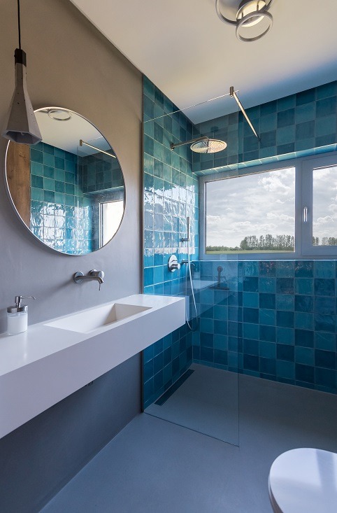 Nowoczesna łazienka z turkusowymi płytkami, minimalistyczną umywalką i okrągłym lustrem. Widok na zewnątrz przez okno prysznica.
