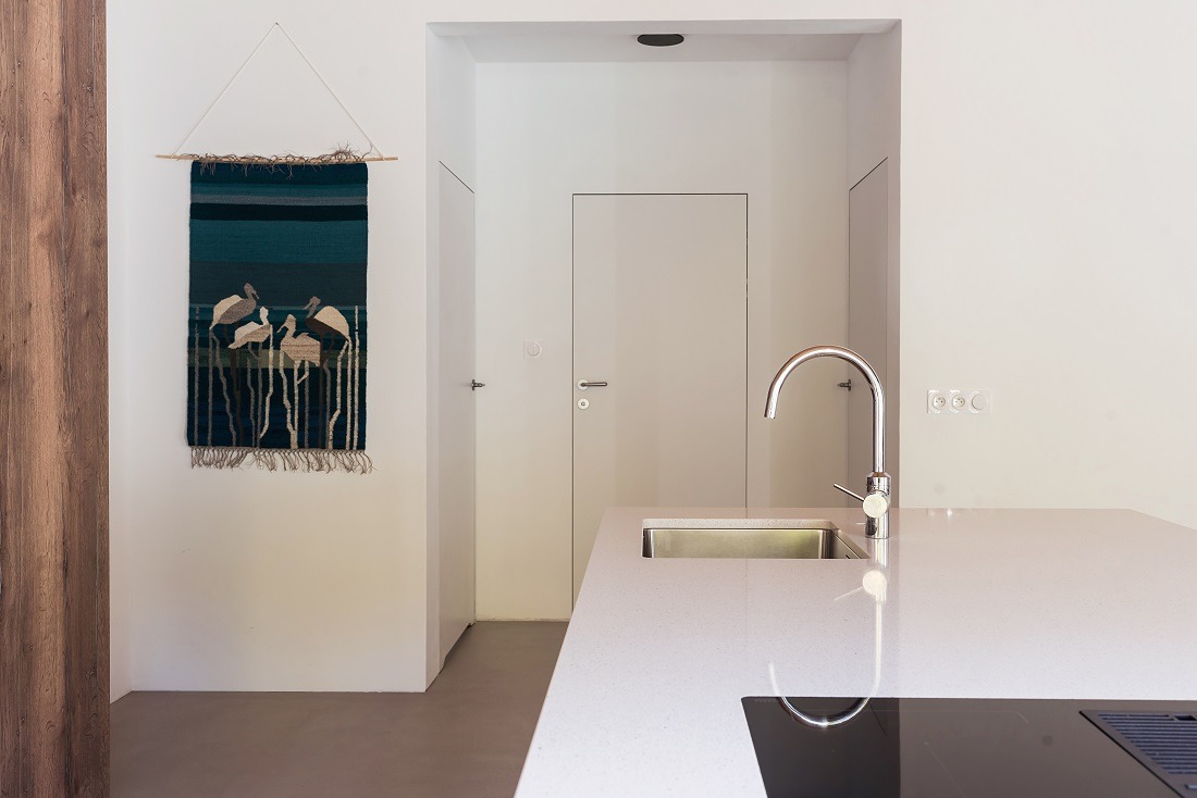 Minimalistyczna kuchnia w Gliwicach: białe ściany, ukryte drzwi, chromowana bateria i dekoracyjny gobelin z motywem ptaków, ukazana w jasnym, naturalnym świetle.