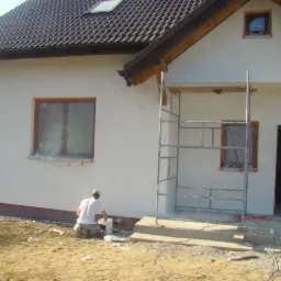 Prace wykończeniowe elewacji domu jednorodzinnego: na kolanach pracownik zabezpiecza cokół budynku, ściany otynkowane na biało, okna zabezpieczone folią, widoczne rusztowanie przy wejściu.