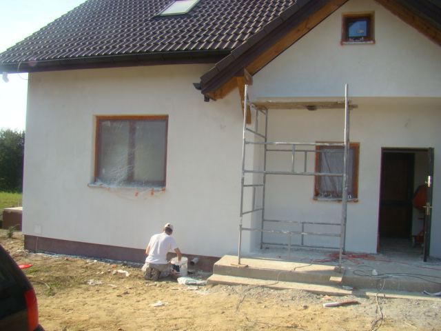 Prace wykończeniowe elewacji domu jednorodzinnego: na kolanach pracownik zabezpiecza cokół budynku, ściany otynkowane na biało, okna zabezpieczone folią, widoczne rusztowanie przy wejściu.