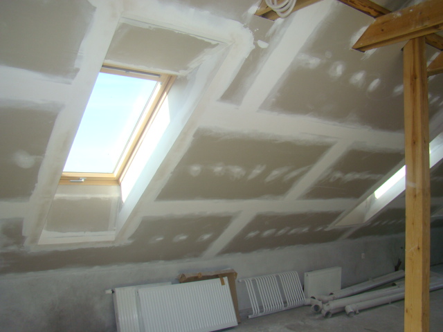 Poddasze w trakcie wykańczania płytami gipsowo-kartonowymi, widoczne okno dachowe i drewniana konstrukcja wspornikowa, grzejniki leżące na podłodze.
