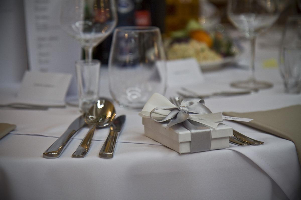 Elegancko nakryty stół z białym obrusem, srebrnymi sztućcami, szklankami i kieliszkami oraz małym, białym pudełkiem z szarą wstążką, prawdopodobnie upominek dla gości.