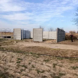 Wznoszenie częściowo ukończonego ogrodzenia z prefabrykowanych paneli betonowych na nierównym, trawiastym terenie; w tle widoczna kobieta z kijem oraz szklarnia.