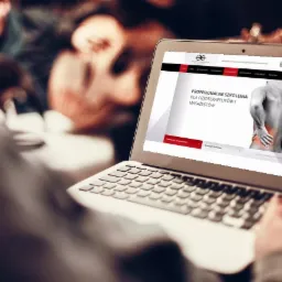 Osoba przegląda na laptopie stronę internetową z ofertą profesjonalnych szkoleń dla fizjoterapeutów i masażystów, widoczny fragment klawiatury i dłoń na touchpadzie, w tle rozmyte sylwetki innych...