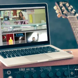 Laptop wyświetlający stronę internetową z muzyką, umieszczony na wzmacniaczu gitarowym obok gitary elektrycznej.