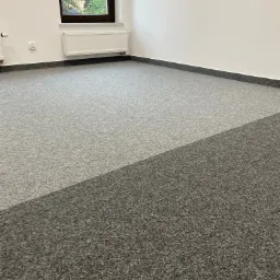 Pokój z dwiema odcieniami szarości wykładziny dywanowej, widok na okno z zielenią na zewnątrz, listwa przypodłogowa w kolorze grafitowym.